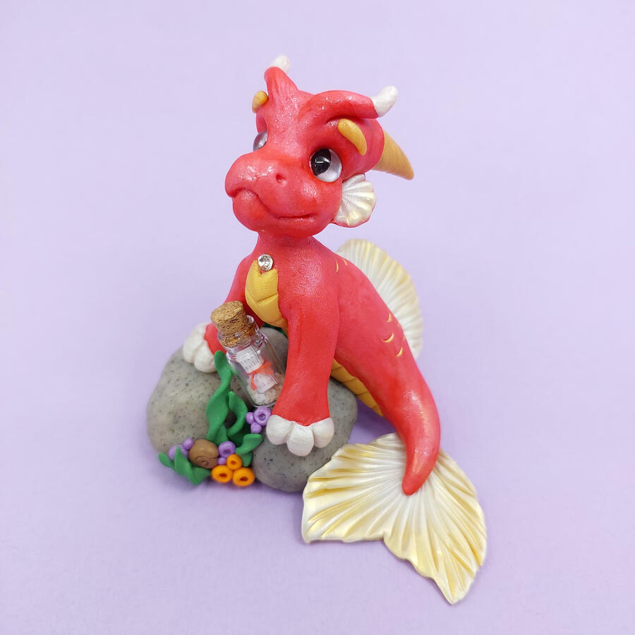 A red clay mermaid dragon propped on a rock covered with seaweed and a glass bottle. The dragon has white and gold fins, and is displayed in front of a purple background.