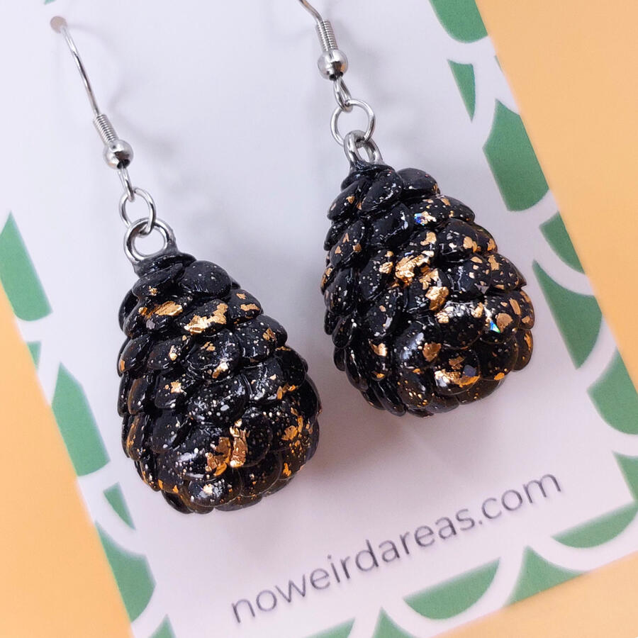 Black and gold scaled egg earrings with silver hooks sitting against a white backing card with a green border. Black and gold scaled egg earrings with silver hooks sitting against a white backing card with a green border.