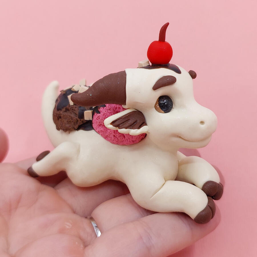 Pale yellow clay dragon lying outstretched on an upturned palm. It has a sculpted cherry on its head, and ice-cream on its back.