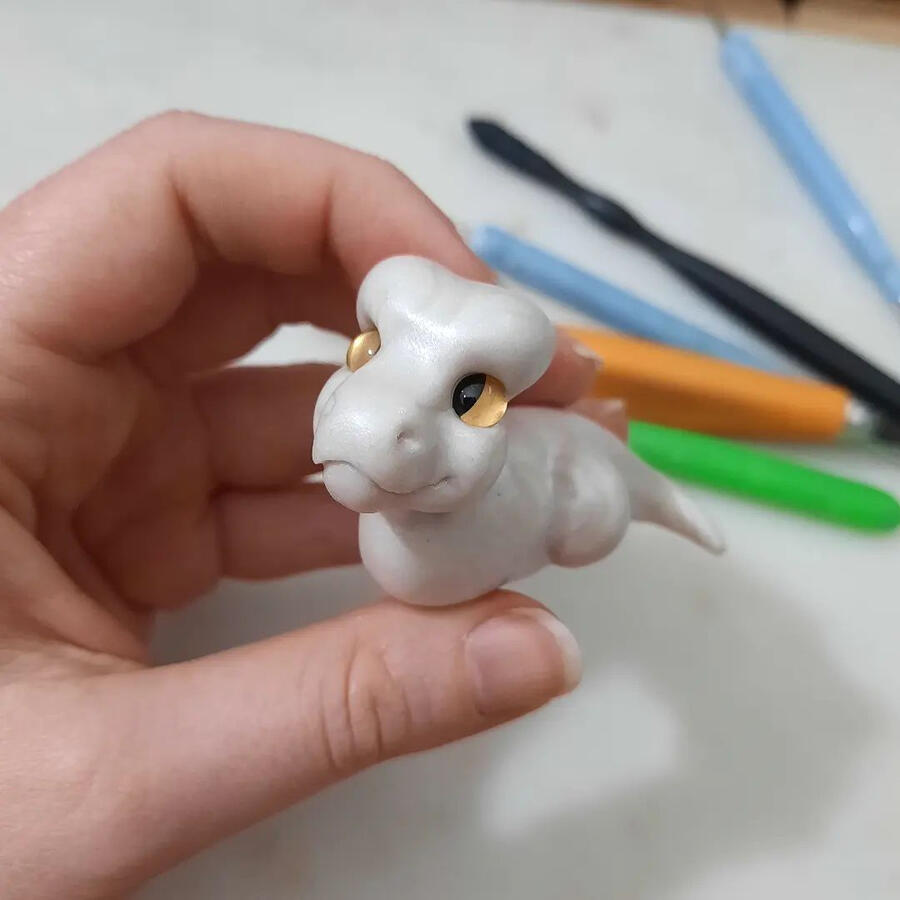 A hand holding a half-finished white clay dragon. The background is slightly blurred, and shows a range of clay tools.