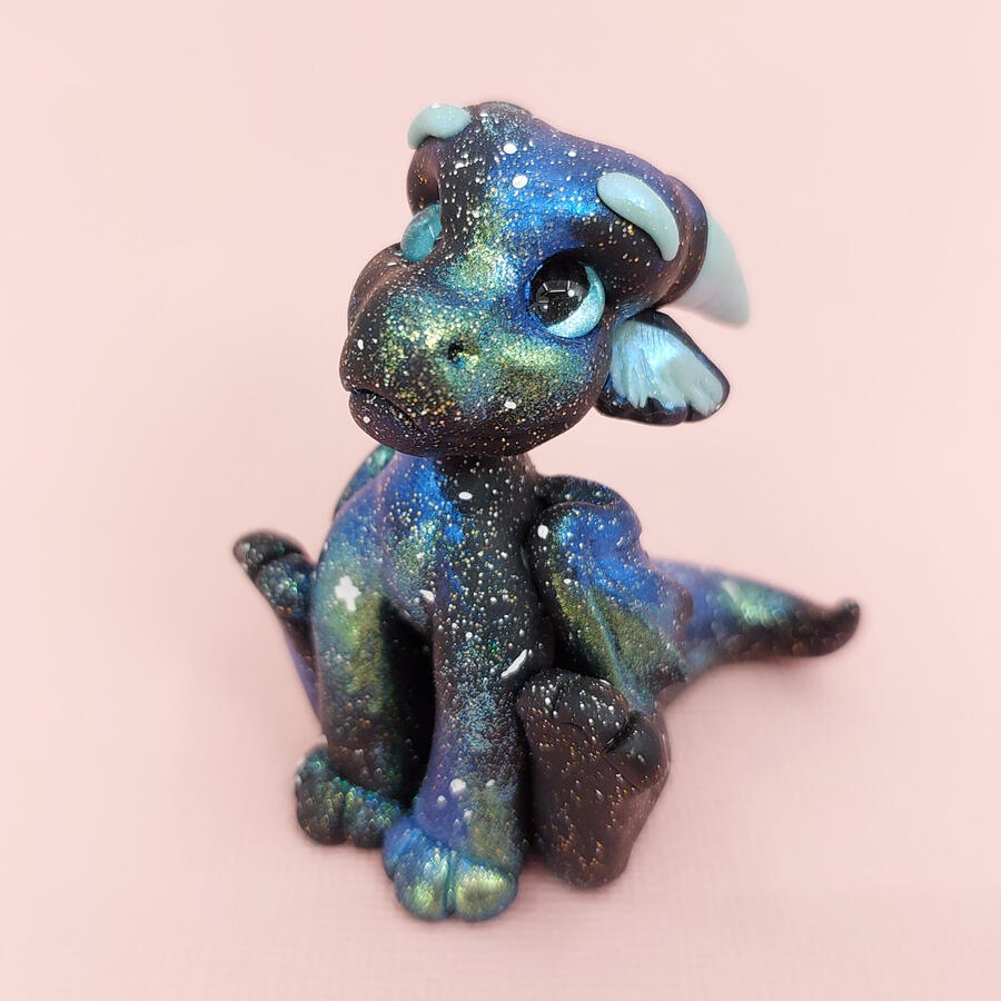 A shiny black and blue clay dragon sits facing slightly away from the camera against a pale peach background.