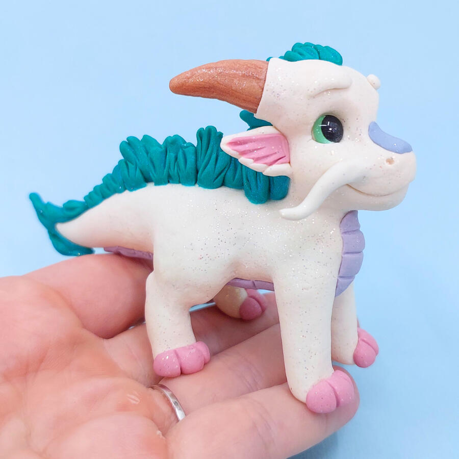 A white clay dragon standing on the fingers of an upturned hand in front of a light blue background.