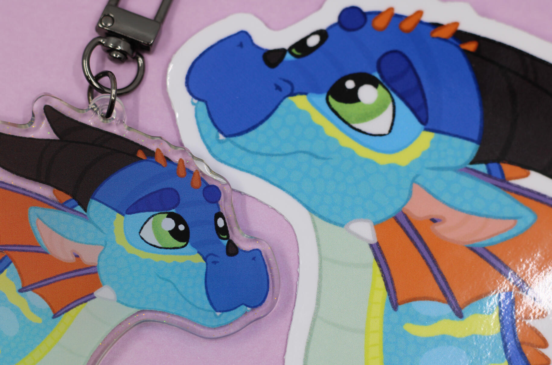 A keychain and sticker of a digital headshot drawing of Glory from Wings of Fire (a blue-green dragon with red-orange frills and black horns).