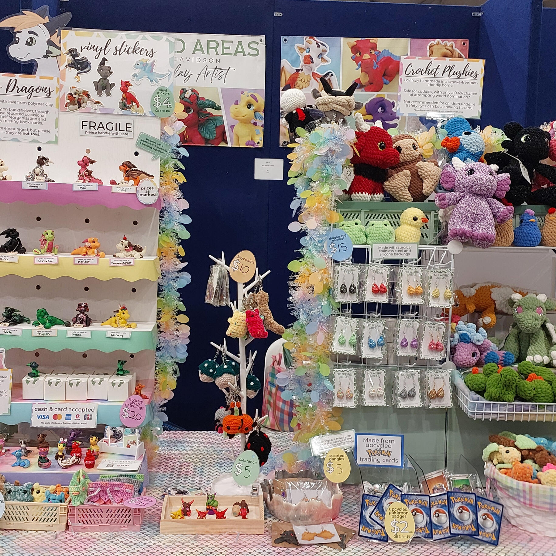 A vendor table display with white shelves on the left stocked with clay dragons, and shelving on the right stocked with plushies. The table is very colourful, and sits against a dark blue backdrop.
